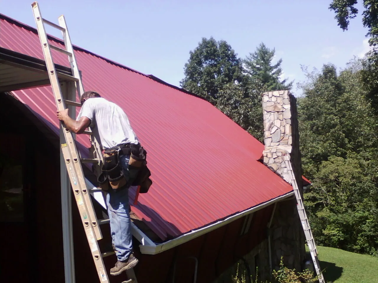 Roofer on a ladder beside a red metal roof and stone chimney — Soffit & Fascia Repair by Roofing Brothers in Jamaica